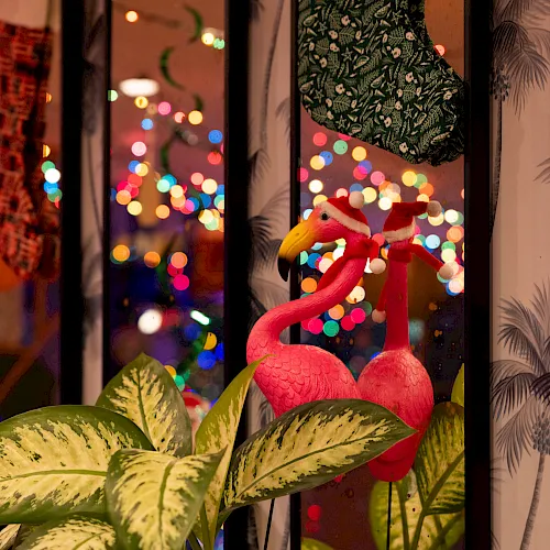 Colorful flamingo ornaments sit by a window with festive bokeh lights outside, giving a cozy holiday glow.