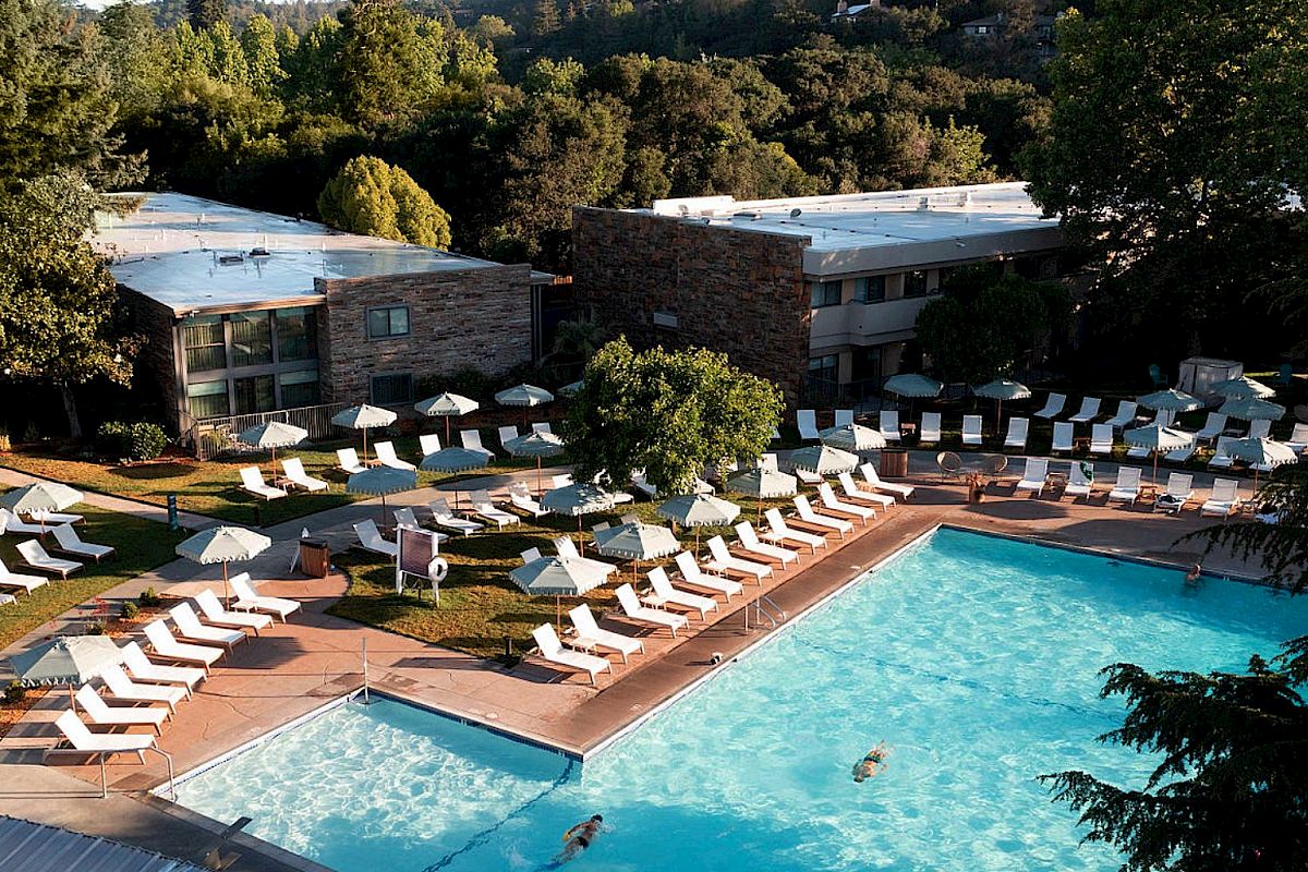 This image shows an outdoor swimming pool area with lounge chairs and umbrellas, surrounded by trees and buildings in the background.