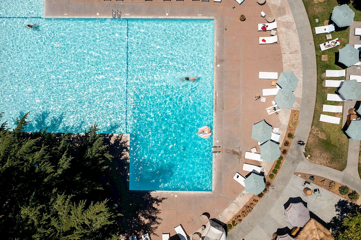 An aerial view of a swimming pool surrounded by lounge chairs and umbrellas, with a few people swimming and relaxing in the water.