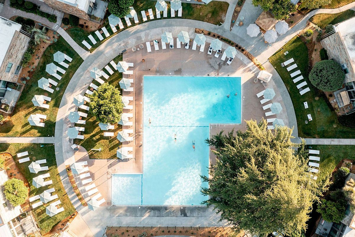 The image shows an aerial view of a large rectangular swimming pool surrounded by numerous lounge chairs and trees, within a landscaped area.