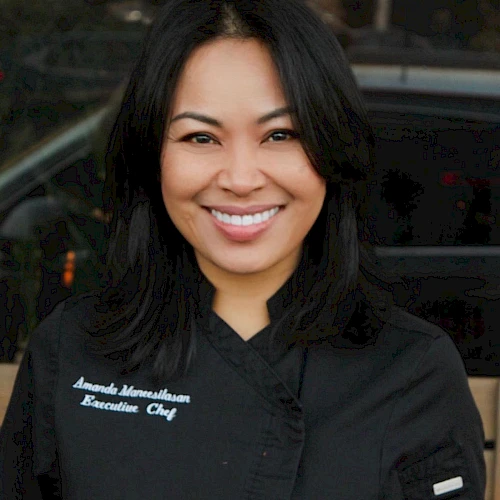 A person wearing a black chef's coat, smiling. The coat has a name and "Executive Chef" written on it.
