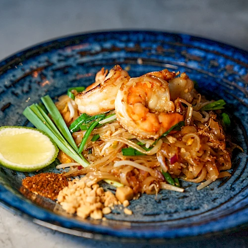 A plate of pad Thai with shrimp, lime wedge, peanuts, and green onions on a blue dish.