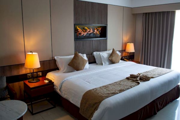 A well-lit bedroom features a large bed with white linens, brown accents, and two nightstands with lamps. A window with curtains is in the background.