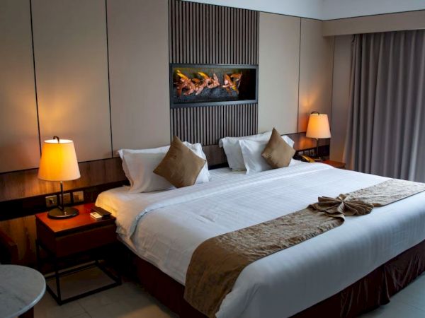 A well-lit bedroom features a large bed with white linens, brown accents, and two nightstands with lamps. A window with curtains is in the background.