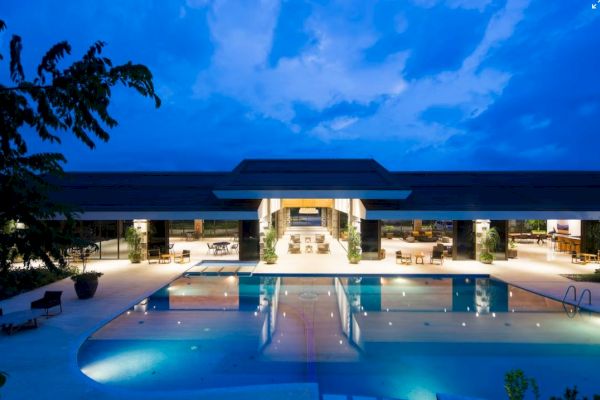 A modern building with outdoor seating and a large illuminated swimming pool at sunset, surrounded by trees and under a dramatic sky.