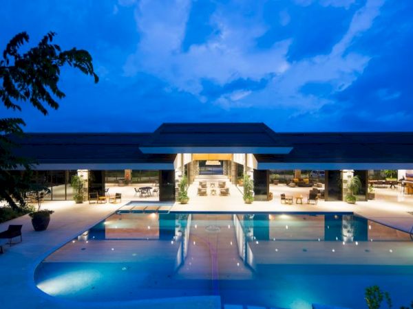 A modern building with outdoor seating and a large illuminated swimming pool at sunset, surrounded by trees and under a dramatic sky.