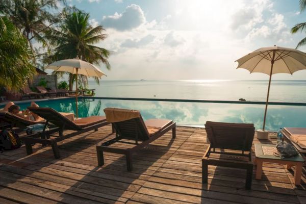 This image shows a tranquil poolside setting with lounge chairs, umbrellas, and a view of the ocean, surrounded by palm trees under a cloudy sky.