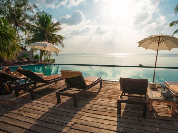 This image shows a tranquil poolside setting with lounge chairs, umbrellas, and a view of the ocean, surrounded by palm trees under a cloudy sky.