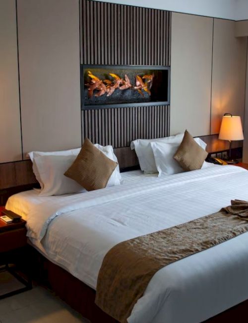 The image shows a cozy hotel room featuring a large bed with white sheets and brown accents, two bedside tables with lamps, and a fireplace.