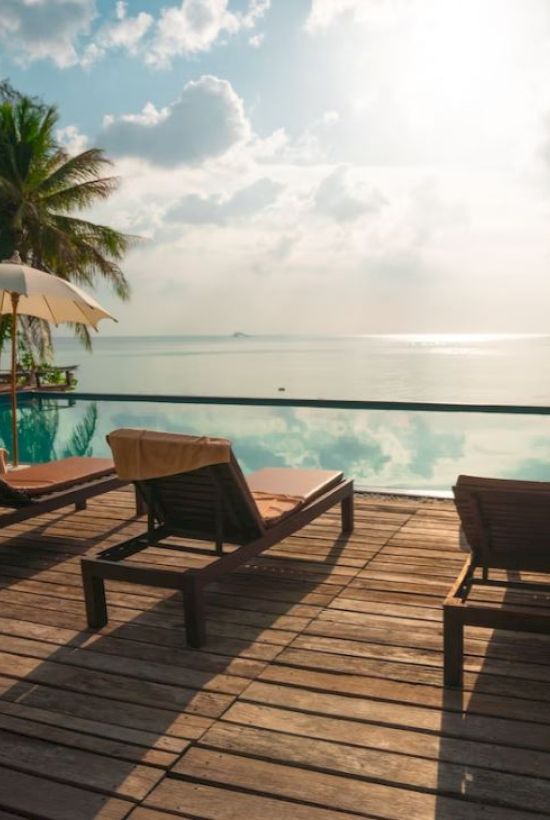 A scenic seaside pool with lounge chairs, umbrellas, palm trees, and a serene ocean view under a partly cloudy sky.