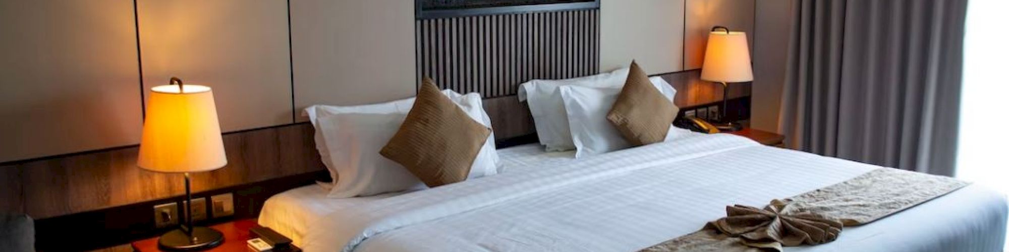 The image shows a modern hotel room with a large bed, side tables with lamps, decorative pillows, and a wall-mounted display. Curtains cover the window.