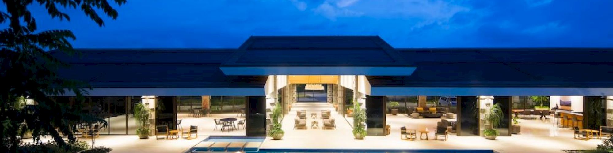A modern building with an illuminated swimming pool in front, surrounded by lounge chairs and tables, under a twilight sky.