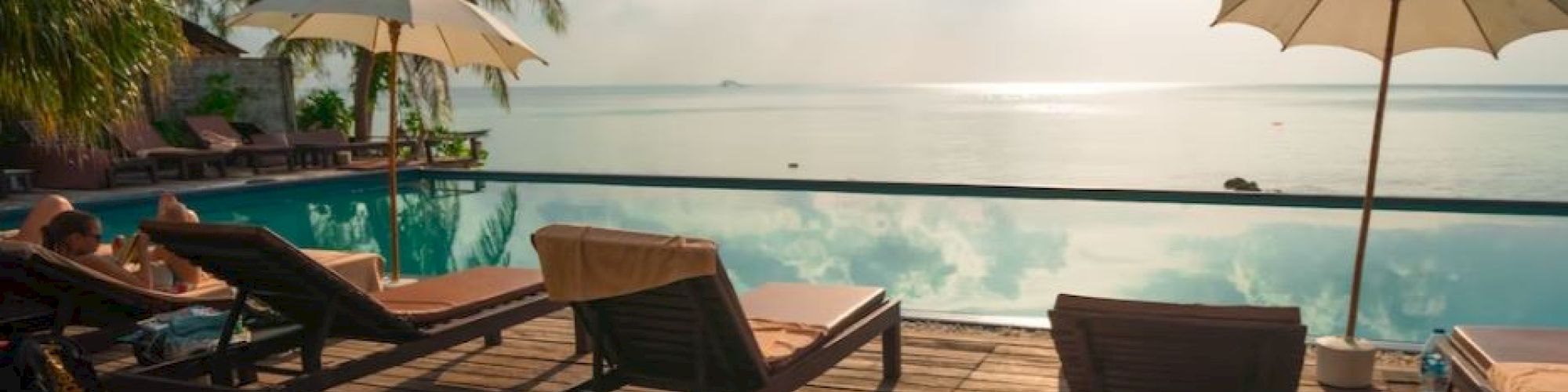 Sun loungers and umbrellas on a wooden deck beside an infinity pool overlooking the ocean, with palm trees and a sunny sky.