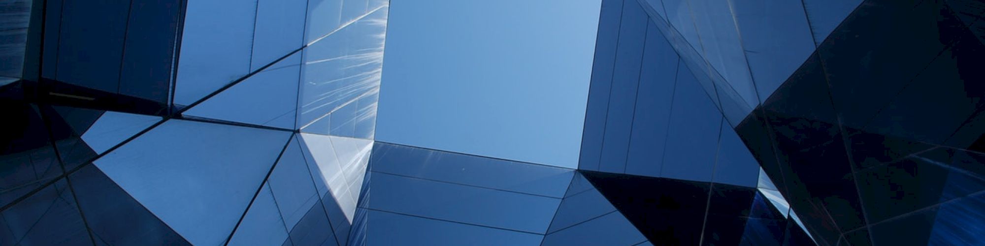 The image shows a geometric arrangement of reflective glass panels forming a prism-like structure with a clear blue sky visible at the center.