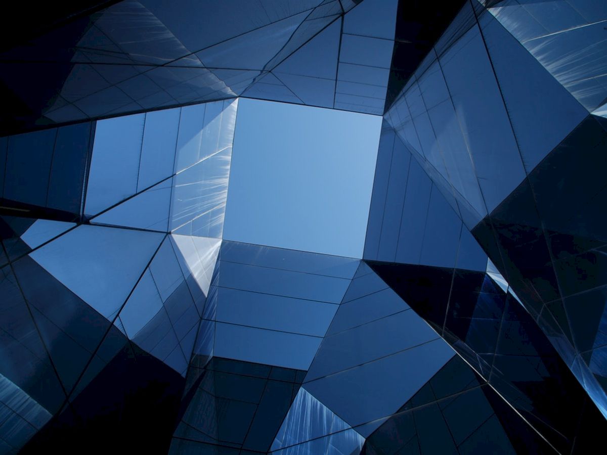 This image features a view of a modern building's geometric, mirrored structure, looking up towards a blue sky through angled reflective surfaces.
