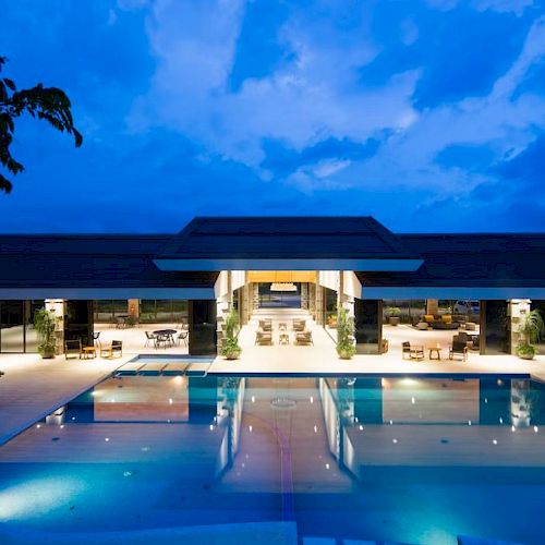 A luxurious, illuminated building with a large, tranquil outdoor pool at dusk, surrounded by comfortable seating areas and greenery.