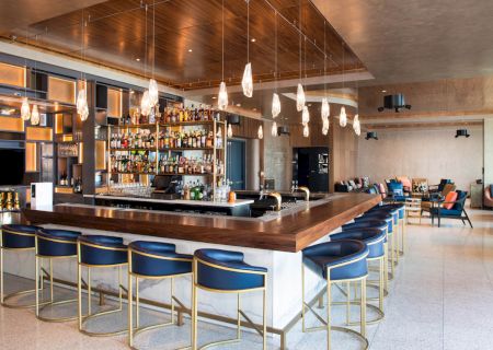 The image shows a modern bar with elegant lighting, a well-stocked liquor shelf, barstools, and a seating area in the background.