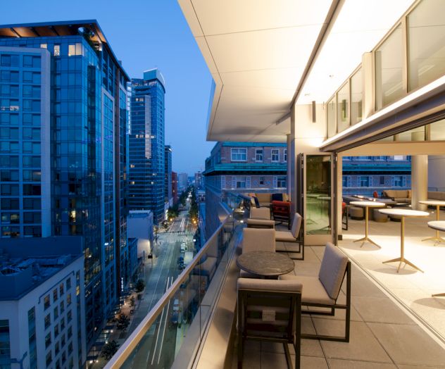 The image shows a modern balcony with chairs and tables overlooking a cityscape with tall buildings at dusk.