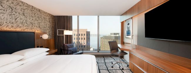 A modern hotel room with a bed, large TV, desk, chair, and windows overlooking a cityscape. The decor is contemporary with wood accents.