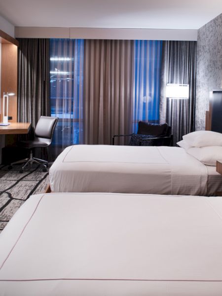 The image shows a modern hotel room with two neatly made beds, a wall-mounted TV, a desk with a chair, and large windows with curtains.