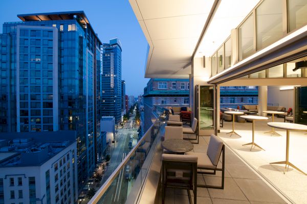 This image shows a modern cityscape with tall buildings as seen from a balcony with seating areas and tables during the evening.