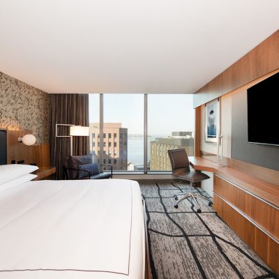 The image shows a hotel room with a large bed, modern decor, a flat-screen TV, a desk and chair, and a window with a city view.
