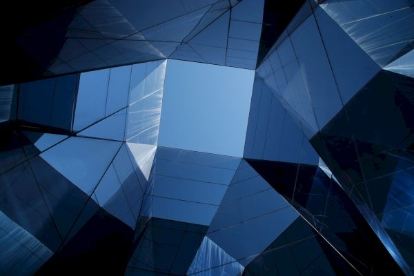 The image shows a geometric arrangement of reflective glass buildings or structures, forming a pattern with a clear blue sky visible in the center.