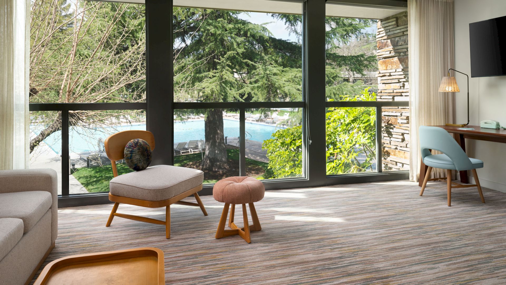 A modern living room with large windows overlooking a lush outdoor scene, featuring comfortable seating and a workspace near the window.