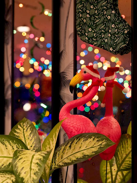 Flamingo decorations sit by a window as colorful city lights blur outside, creating a festive, cozy night scene.