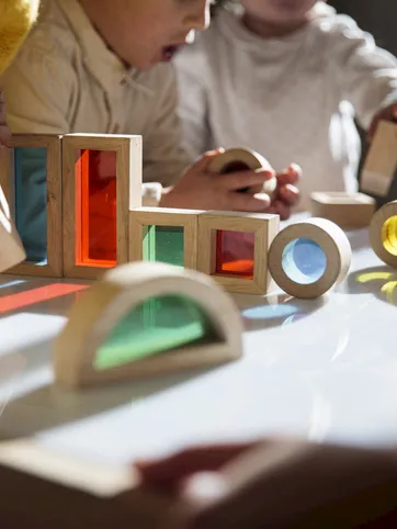 Kids play with colorful wooden block toys, glass-like panels, and spinning pieces on a table, laughing together.