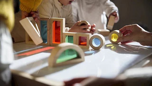Kids play with colorful wooden block toys, glass-like panels, and spinning pieces on a table, laughing together.