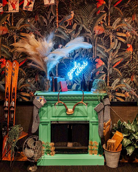 A wild decorated room with tropical bird wallpaper, glowing blue neon, skis, plants, and a feathered centerpiece by a green bar counter.