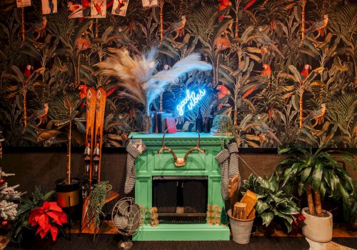 A wild decorated room with tropical bird wallpaper, glowing blue neon, skis, plants, and a feathered centerpiece by a green bar counter.