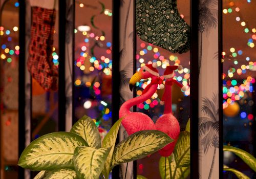 Colorful flamingo ornaments sit by a window with festive bokeh lights outside, giving a cozy holiday glow.