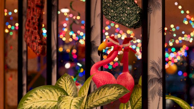 Colorful flamingo ornaments sit by a window with festive bokeh lights outside, giving a cozy holiday glow.