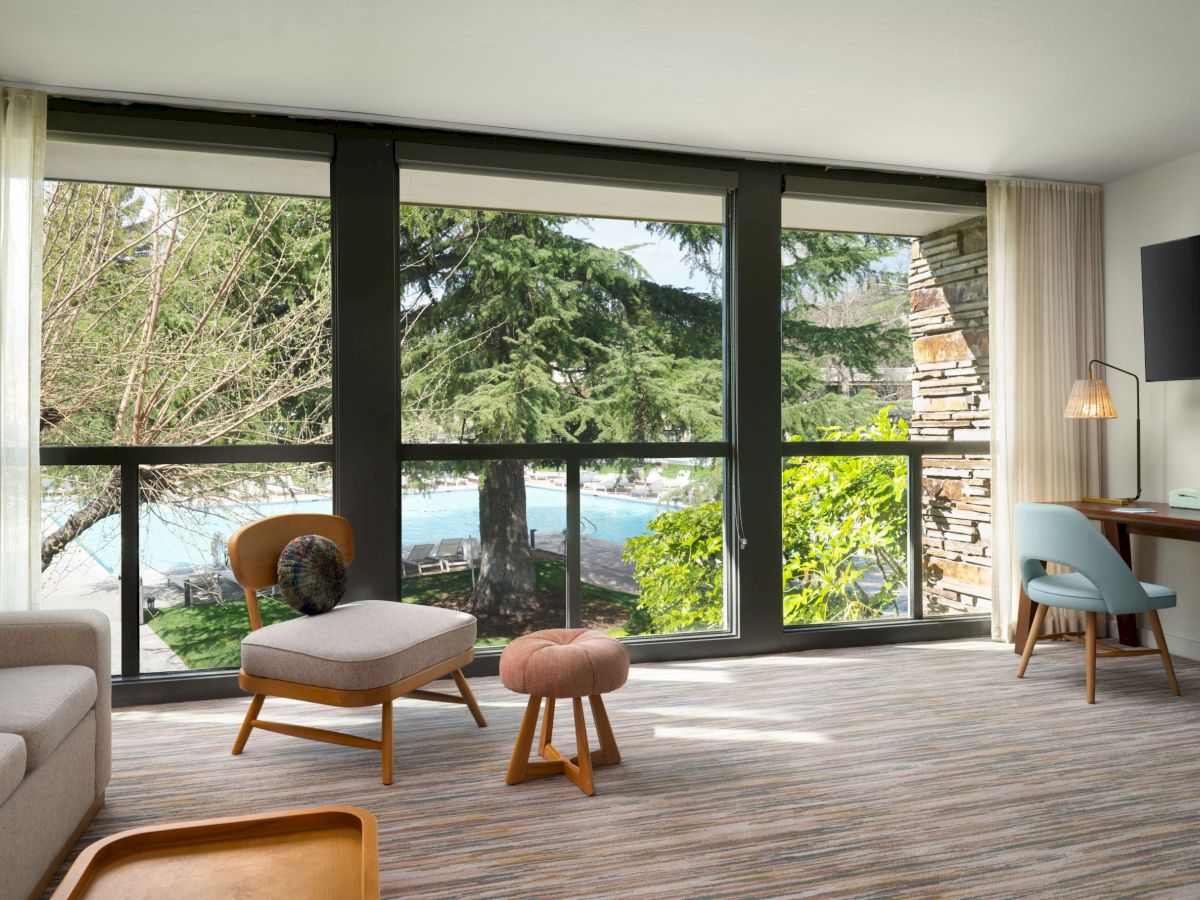 A modern living room features large windows showing a scenic outdoor view, with minimalist furniture, a desk, and a wall-mounted TV.