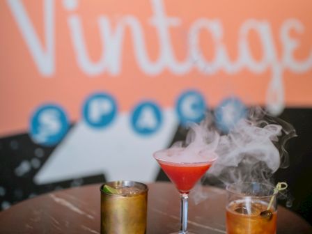 The image shows three cocktails on a table with a vintage-themed background displaying the word "Vintage." The cocktails appear to have different colors and garnishes.