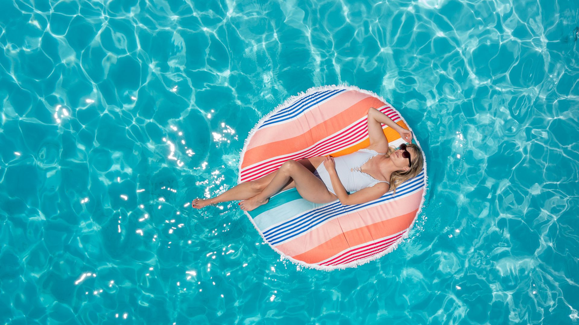 A person is relaxing on a colorful float in a swimming pool, basking in the sun. They are wearing sunglasses and enjoying the clear blue water.