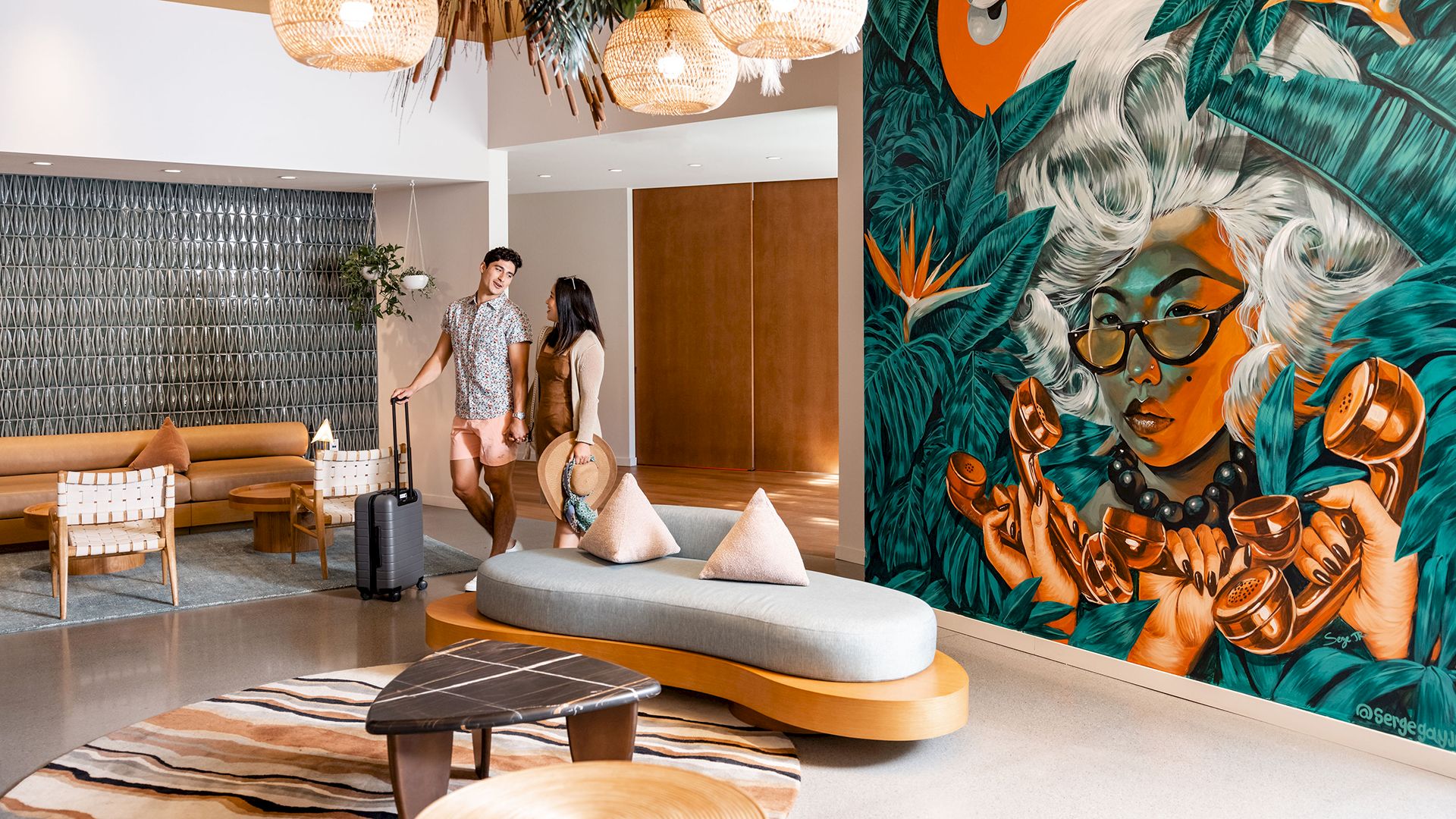 A modern hotel lobby with a vibrant mural, stylish furniture, and two people near the reception. They have suitcases and are conversing.