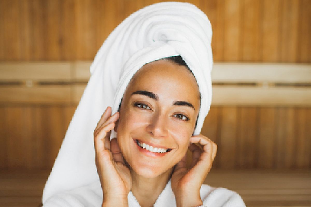 A person in a white bathrobe and towel, smiling in a wooden sauna room, hands near face.