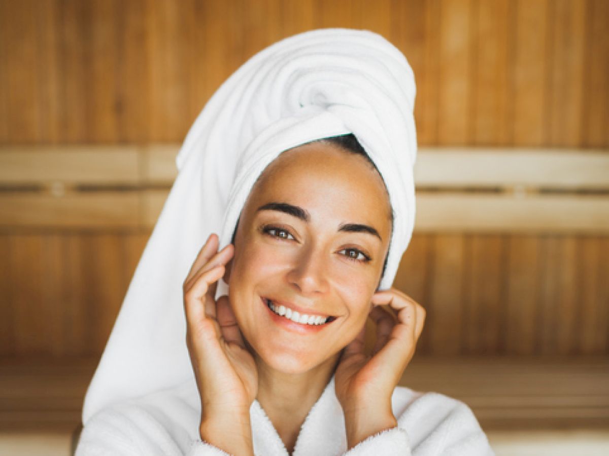 A person in a white bathrobe and towel, smiling in a wooden sauna room, hands near face.