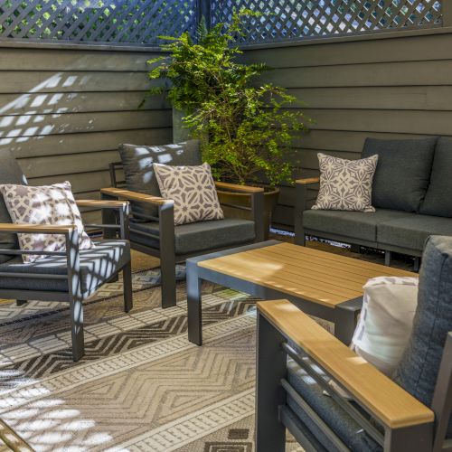 A modern outdoor patio with cushioned seating, a coffee table, and potted plants; sunny deck, light wood furniture, and a grill setup.