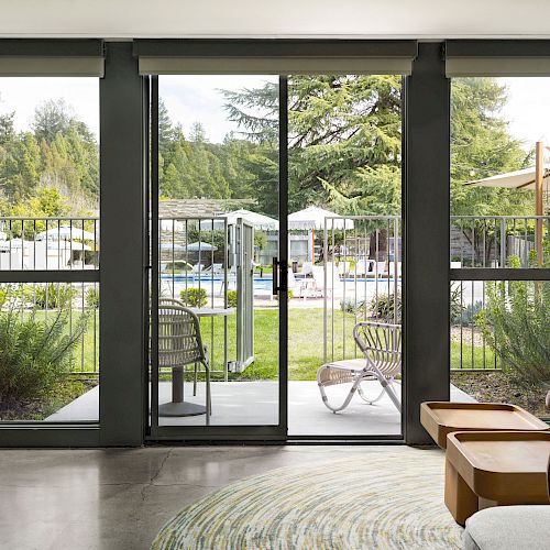 The image shows an interior view of a living room with large sliding glass doors leading to an outdoor pool area with chairs and umbrellas.