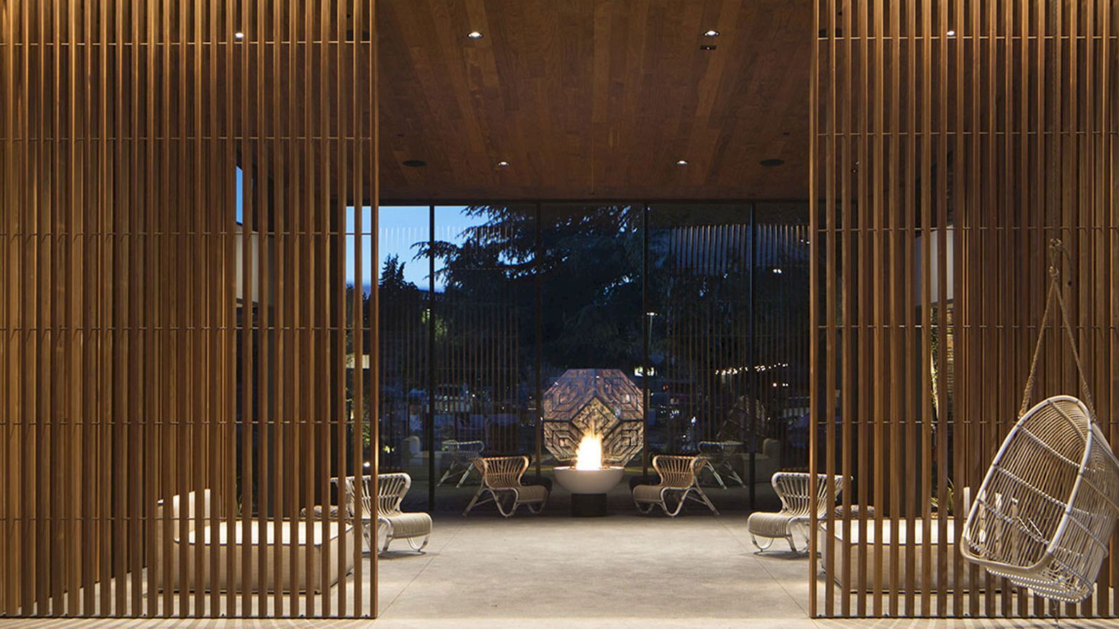 The image shows a modern patio area with wooden vertical slats, hanging chairs, lounge seating, and a central fireplace, all under ambient evening lighting.