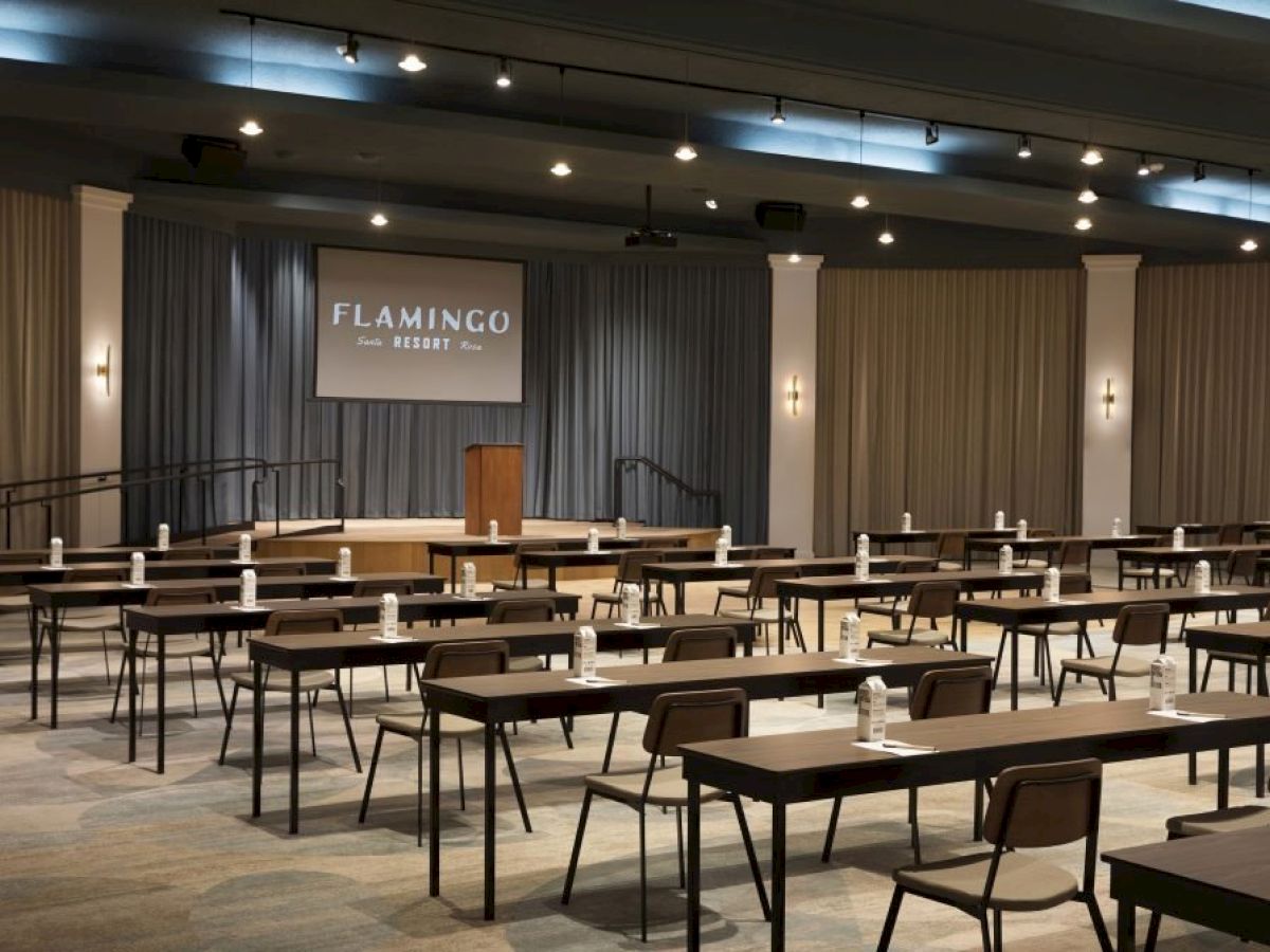 The image shows a conference room at Flamingo Resort with rows of tables and chairs, a stage, a podium, and a projection screen.