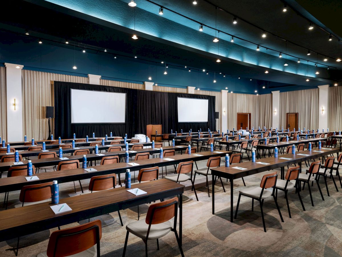 A conference room with rows of tables and chairs facing two large screens, each setup with water bottles and notepads on the tables.
