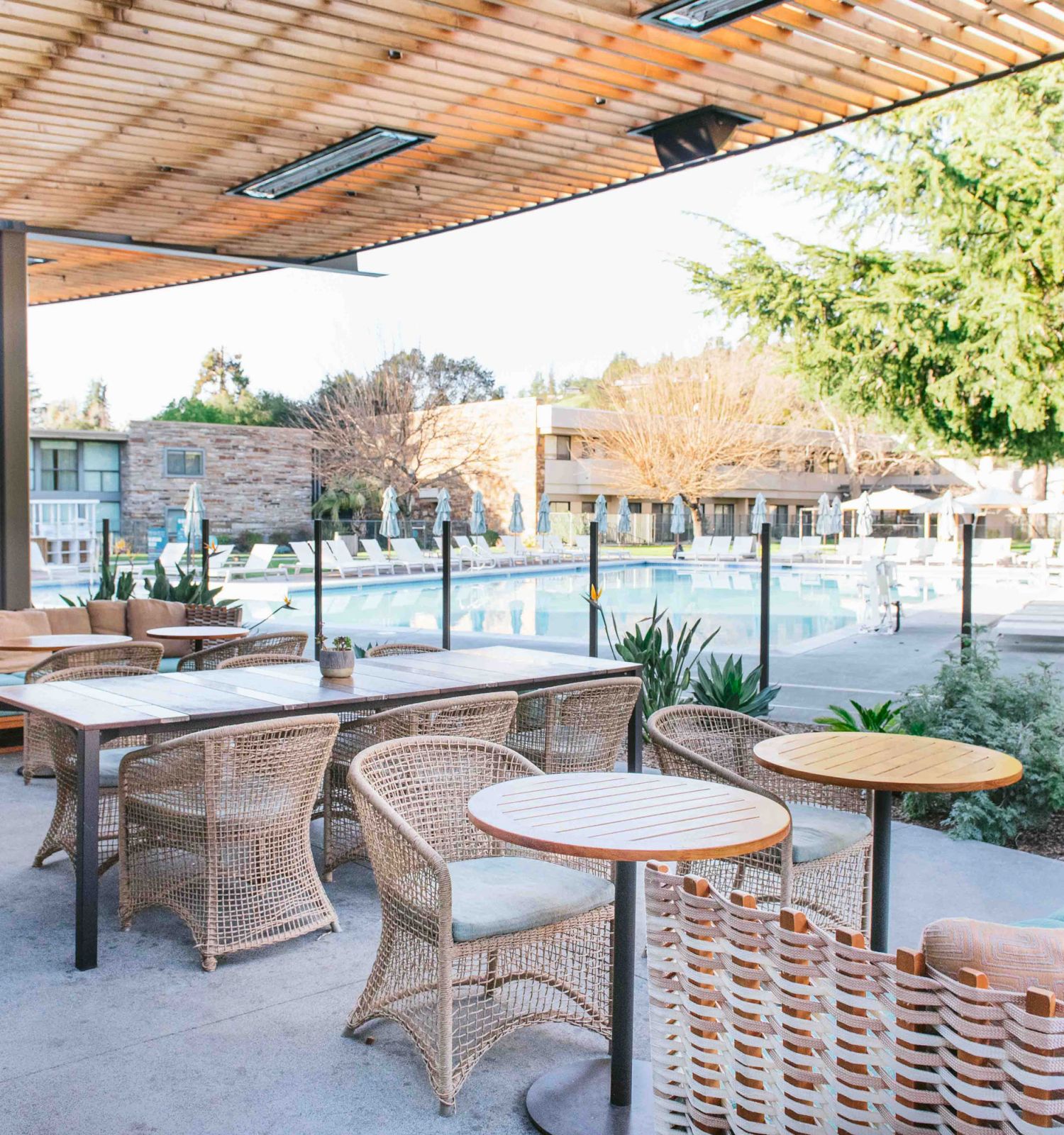 An outdoor seating area with wicker furniture and tables, overlooking a swimming pool and surrounded by trees.