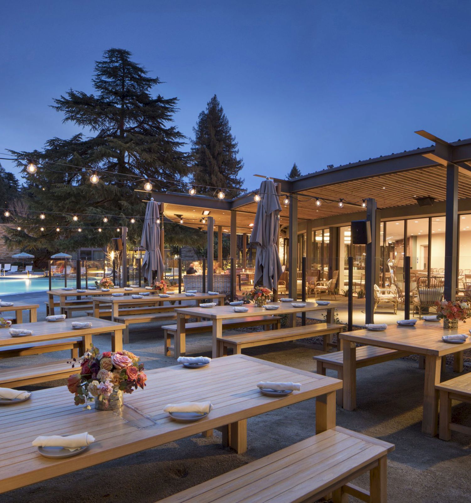 An outdoor dining area with wooden tables and benches is decorated with flowers and string lights, under a pergola, beside a pool at twilight.