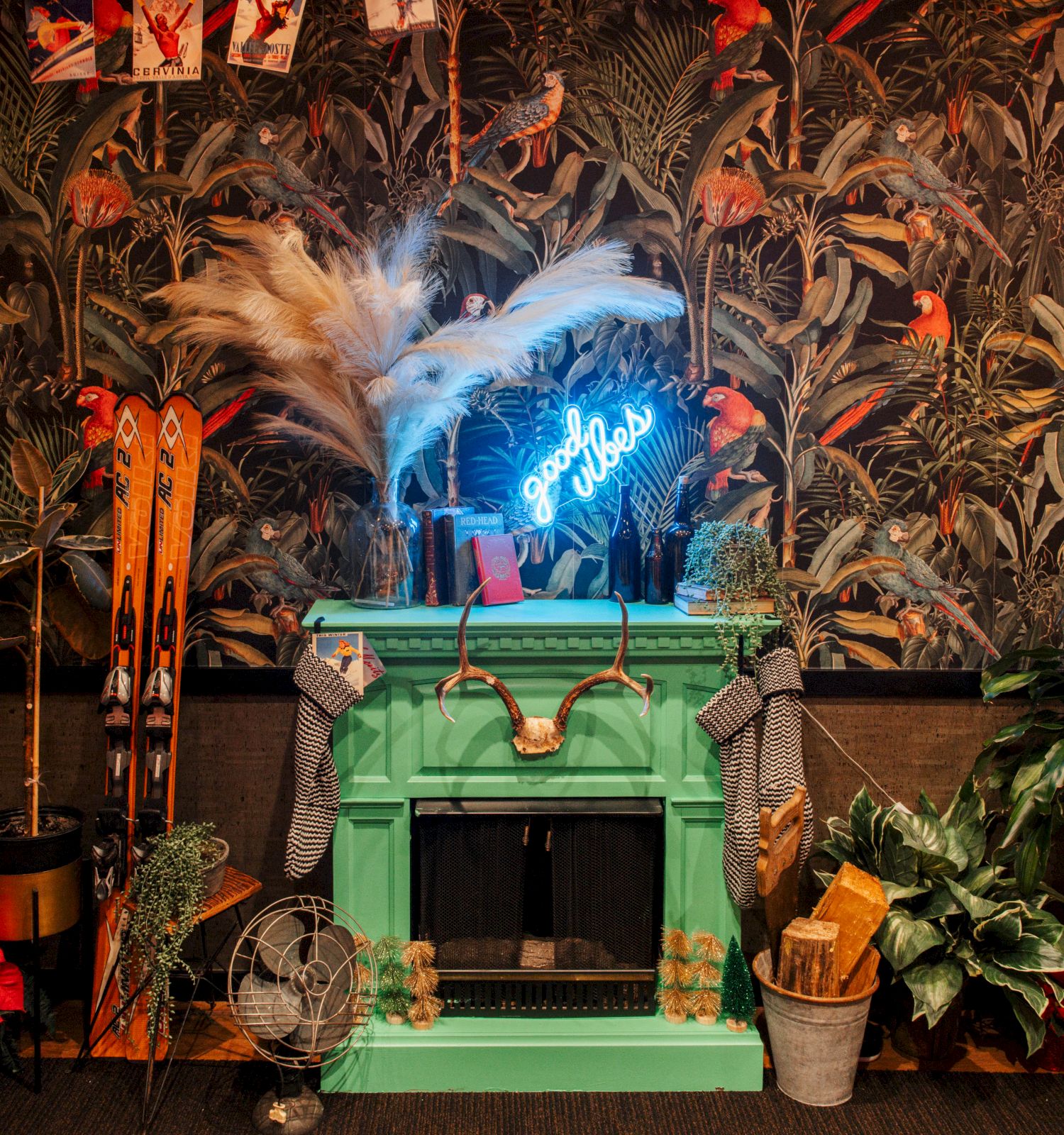 A cozy fireplace with a neon sign, surrounded by plants, skis, wood, fan, and decor against a tropical wallpaper.