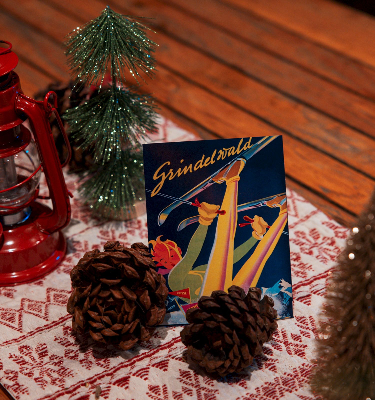 A festive table setup with a red lantern, pinecones, small decorated trees, and a "Grindelwald" ski poster on a patterned runner.